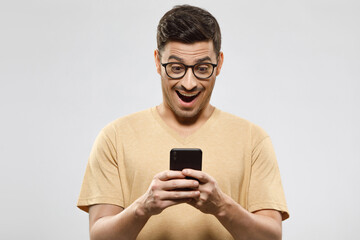 Young handsome guy dressed in beige t-shirt, wearing eyeglasses, holding cellphone in hands, looking at screen with excited face, isolated on gray background
