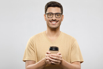 Fototapeta premium Young man in t-shirt and eyeglasses standing with takeaway coffee to go cup in hands, ready to enjoy his break from work, isolated on gray background