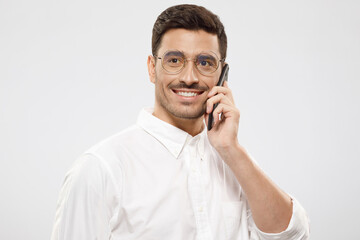 Portrait of young positive handsome businessman in shirt and glasses, answering phone call, looking aside with smile, isolated on gray background