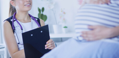Young woman doctor with stethoscope and tablet speaking with pregnant woman at hospital.