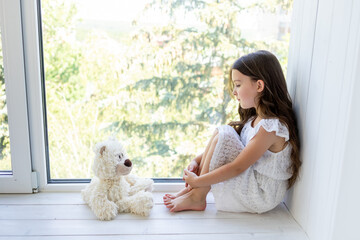a charming little girl 5-6 years old looks at a Teddy bear sitting by the window