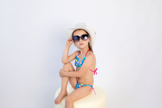 Cute little girl in black sunglasses and a blue bikini is sitting on the floor , turning sideways to the camera-Isolated on white background