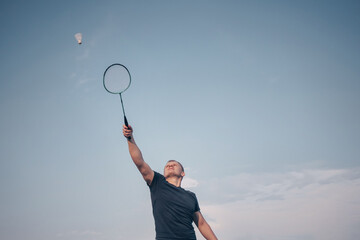 Young Caucasian man plays badminton on a background of blue sky. The concept of an amateur game of badminton, outdoor activities. Copyspace, bottom view..