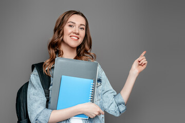 Student girl holds folders, books, notebooks, notebooks in hands and points a finger at copy space isolated on gray wall background