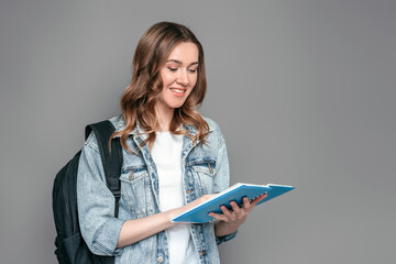 Student girl holding a notebook with homework in her hands reading and smiling isolated over grey...