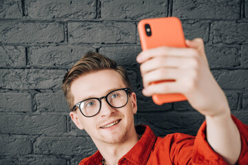 Portrait young man in red shirt and glasses taking selfie photo on cellphone on a background of black brick wall. Copy, empty space for text