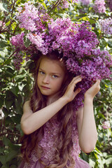 Caucasian girl child seven years old in a purple dress stands in nature with a wreath of lilacs