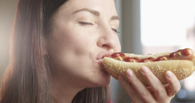 Hungry Woman Eating Hot Dog Enjoying the Taste Indoors with Sun Shining a Close up Shot on Red Camera