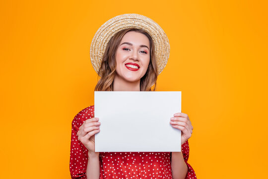 Portrait of a girl in a red dress, a straw hat with red lips smiling and holding in her hands an A4 format poster isolated over orange background, mockup for greeting card text