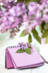 an inflorescence of lilac with leaves lying on a lilac colored Notepad and a blurry bouquet of lilac in a white vase