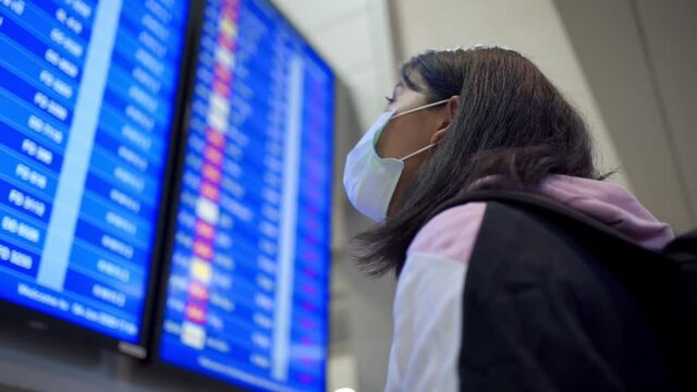 Asian female passenger wear protective mask standing in front of airline schedule time board, at airport terminal, covid-19 pandemic, new normal, flight time schedule, close up look up, lone traveler