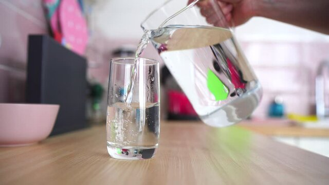Woman's hand pouring fresh pure water from jug a glass on the kitchen table, health and diet concept.