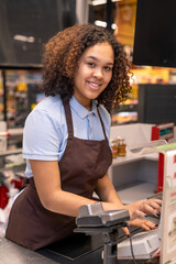 Happy young mixed-race cashier with dark wavy hair pressing button on cashbox
