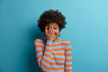 Fototapeta na wymiar Sincere emotions and happiness concept. Shy feminine girl covers face, looks sensually, feels happy and upbeat, has positive mood, dressed in casual striped jumper, isolated over blue background