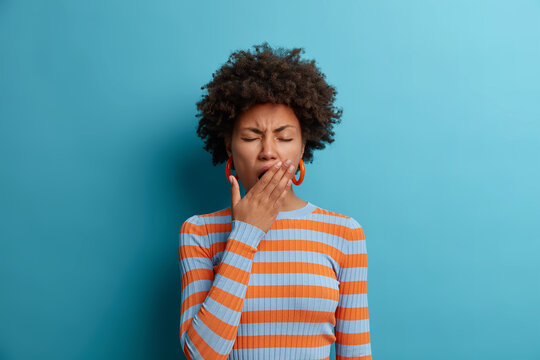 Tired Exhausted Afro American Woman Feels Bad After Hard Day At Work, Covers Mouth And Has Sleepy Expression, Wears Casual Striped Jumper, Isolated On Blue Background. Restless And Sleepiness Concept