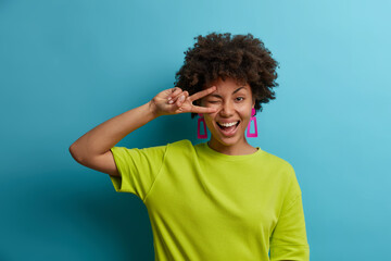Charming friendly African American woman makes peace sign over eye, smiles broadly and feels positive emotions, sends good vibes, wears bright green t shirt. People, emotions, summertime concept