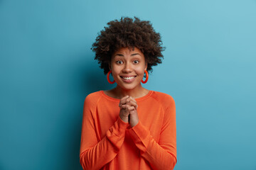 Fototapeta na wymiar Beautiful positive African American woman has imploring look, keeps hands pressed together, hopes for better, wears orange sweater and earrrings, asks for help, pleads for something, poses indoor