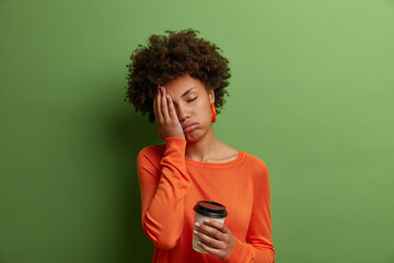 Fototapeta na wymiar Sleepy exhausted dark skinned curly woman holds cup of coffee, cannot wake up and go to work, had sleepless night, wears casual orange jumper, has sad expression, isolated on green background