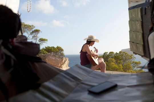 Wanderlust concept of a woman with cowboy look traveling in a van, the girl is wearing a hat and the evening light shines in the sky. Pretty woman play the guitar in seascape of Majorca . Van trip.
