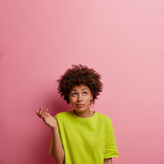 Isolated shot of ethnic curly woman shrugs shoulders and stands hesitant, raises palm, looks above confused, wears green t shirt, isolated on pink background, empty space for your information