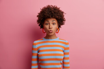 Lovely woman with fluffy frizzy hair, keeps lips rounded, sends air kiss, flirts and demonstrates love, has romantic mood, wears casual striped jumper, isolated over pink background. Sensual kissing