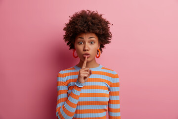 Surprised woman tells secret, holds finger on lips in silence gesture, gossips about something, asks to stay quiet, wears striped sweater, poses against pink background. Hush, dont talk, keep calm