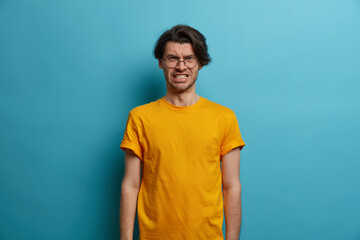 Irritated European hipster clenches teeth with annoyance, looks at camera with mad face, wears casual yellow t shirt and transparent glasses, expresses negative emotions. Furious expression.