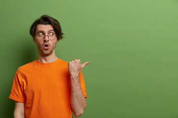 Photo of shocked impressed guy points away with thumb, shows copy space on green background,...