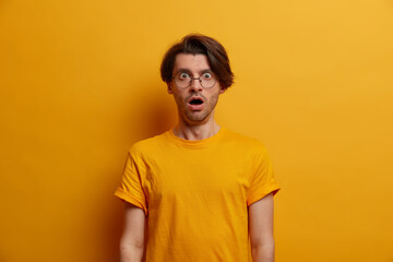 Portrait of astonished brunet man gasps from great wonder, keeps mouth widely opened, reacts on shocking news, stands speechless, wears optical glasses and t shirt, yellow background. Monochrome