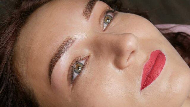 Portrait of a beautiful young woman with permanent make up microblading with eyebrow tattoo and lips with red lipstick lies on a couch in a beauty salon after the procedure.