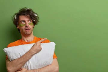 Horizontal shot of surprised speechless man points away with index finger, shows blank space for commercial, holds pillow, awakes and applies patches under eyes, isolated on green background