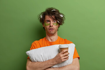 Portrait of shocked man awakes in morning, drinks coffee to go, embraces pillow, stands indoor...