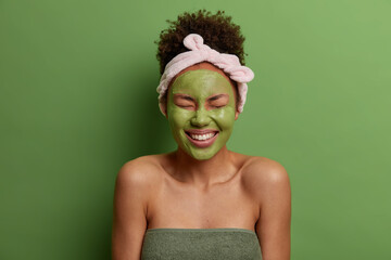 Young overjoyed woman applies natural clay mask for reducing acnes, smiles broadly, has perfect white teeth, has combed curly hair, headband, shows bare shoulders, isolated on green background
