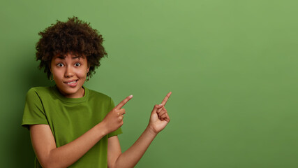 Puzzled dark skinned woman with Afro hairstyle bites lips, points at upper right corner, demonstrates decreased price or special offer, dressed in casual green t shirt, poses indoor. Advertisement