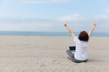 Fototapeta na wymiar Happy freelance woman raised her hands up while sitting on the empty beach / sand with laptop. 