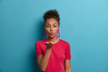 Portrait of romantic African American woman expresses love to someone, keeps lips rounded and palm towards camera, flirts with boyfriend, sends air kiss, dressed in red t shirt, isolated on blue