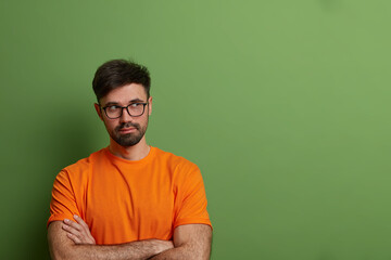 Thoughtful male model with serious expression, stands with crossed hands and looks away, thinks what to do or makes plans, has thick stubble, isolated on green background, blank space aside.