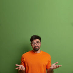 Vertical shot of questioned clueless man shrugs shoulders and raises palms, has doubtful expression, concentrated above, hesitates what to choose, poses against green wall, empty space upwards