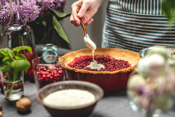 Cooking homemade cranberry pie. Female pastry chef is adding of sour cream with spoon to a baked tart with lingonberries