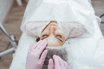 Beautician doctor in pink gloves is applying facial cleansing foam on woman's face massaging skin in cosmetology clinic.
