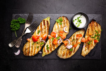 Grilled eggplant slices, garnished with fresh herbs, on black background, top view