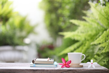 White coffee cup and notebooks with flowers on brown wooden table at outdoor lush scenery 