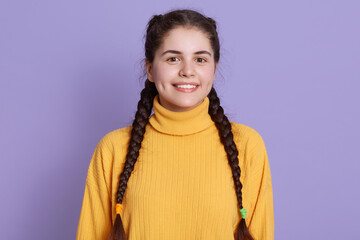 Charming dimpled girl with two pigtails posing against lilac wall, wearing yellow sweater, looking...