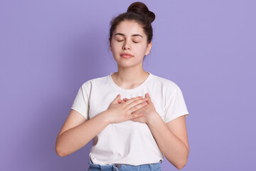 Young lady wearing white t shirt keeps hands on chest and standing with closed eyes, female with...