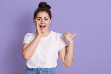 Excited smiling happy woman touching her cheek with palm and pointing aside with thumb, showing something amazement, keeps mouth opened, wearing white t shirt.