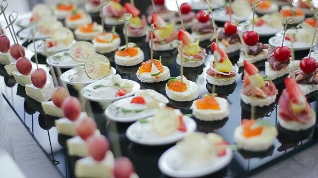 Appetizing and delicious wedding snacks on skewers for guests, such as strawberries, cherries, brie cheese, red caviar, pineapple, lemon on a black table tray in the morning at the banquet hall.