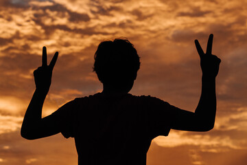 Silhouette of a kid raising his arms and doing peace sign with his hand in front of the beautiful...