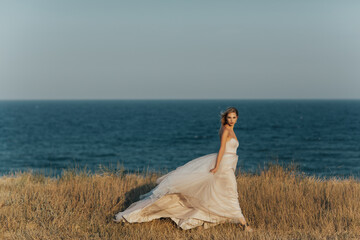 Fototapeta na wymiar Young beautiful girl in a long rose pale dress walks along the beach against the background of the blue sea. Bride walking along sea coast wearing beautiful wedding dress. Copy space.