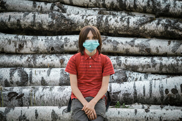 A boy in a medical mask stands near birch logs