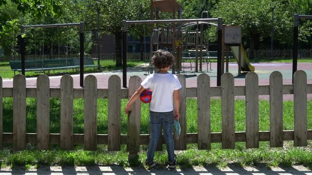 Europe, Italy , Milan - sad boy with mask and ball arrives to the park and the play area is closed and empty of children due COVID19 Coronavirus quarantine home lockdown